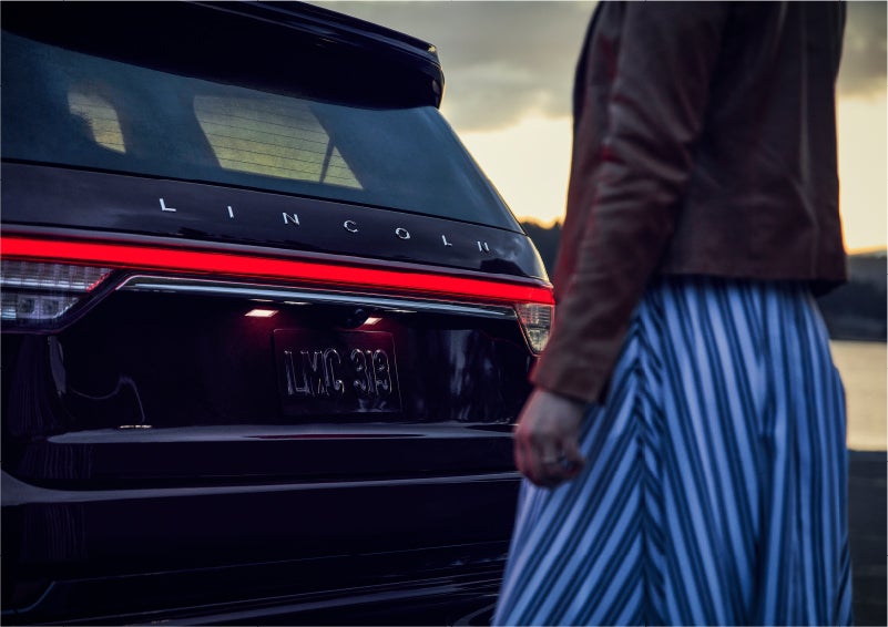 A person is shown near the rear of a 2023 Lincoln Aviator® SUV as the Lincoln Embrace illuminates the rear lights | Casa Lincoln in El Paso TX